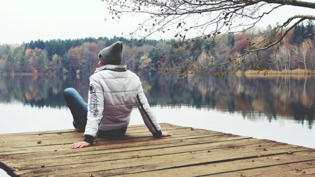 A person sitting on a wooden dock by a calm lake, wearing a white jacket and grey beanie, surrounded by autumn foliage and trees reflecting in the water.