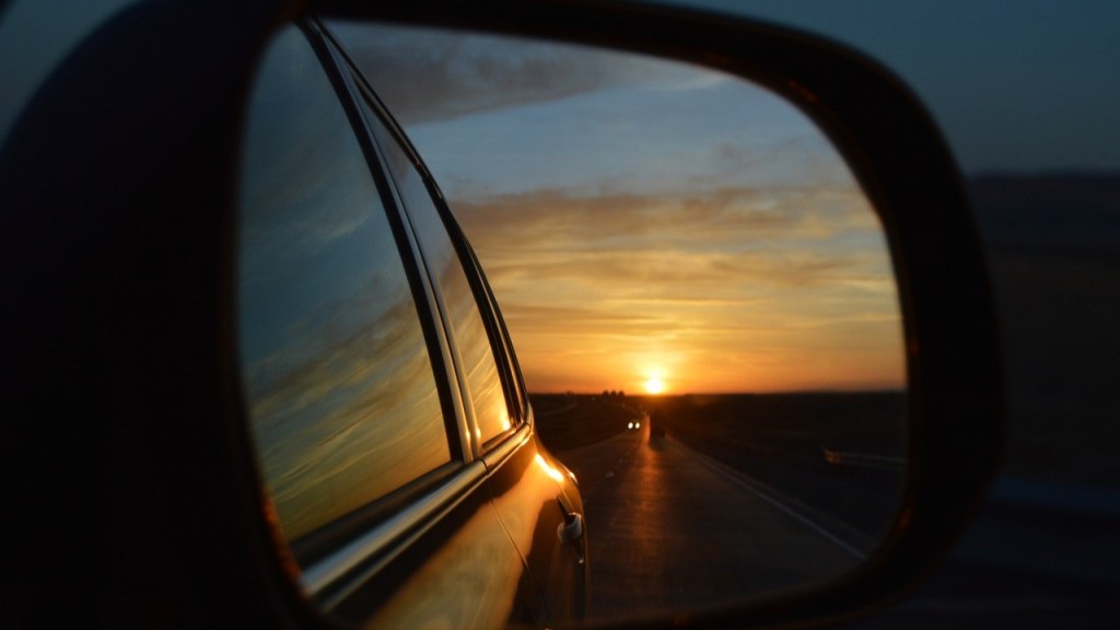 Side mirror view of a car reflecting a sunset over a road, with two vehicles in the distance.