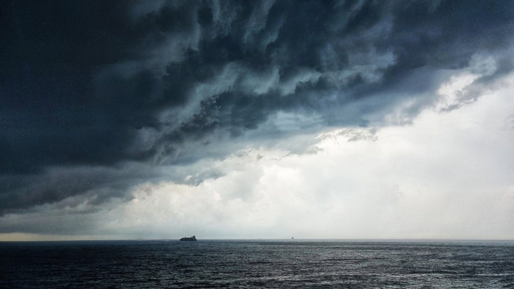 A stormy seascape with dark clouds gathering above the ocean, featuring a distant cargo ship and a faint outline of another vessel on the horizon.