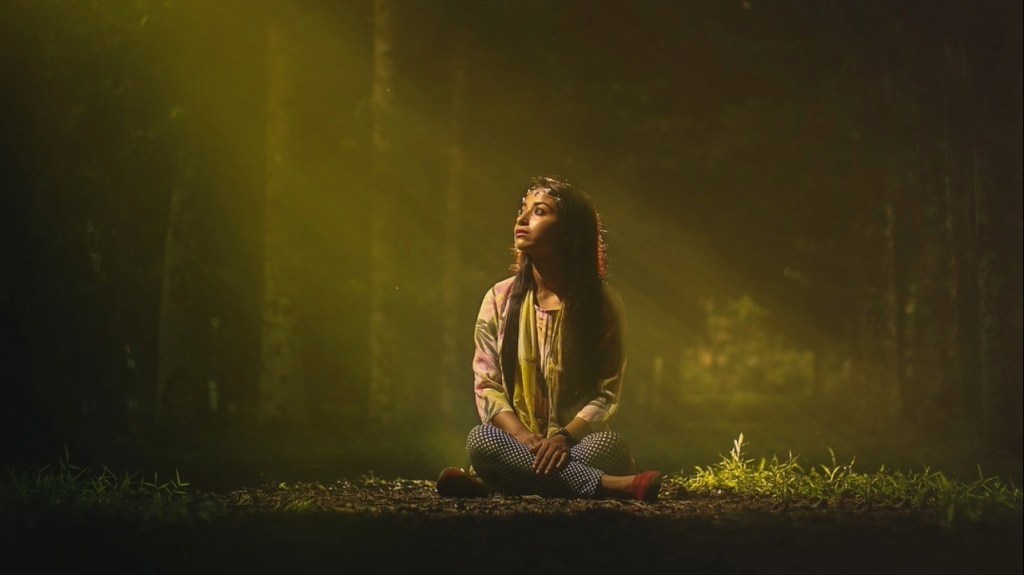 A woman sitting cross-legged on the ground in a forest, surrounded by soft, golden light filtering through the trees.