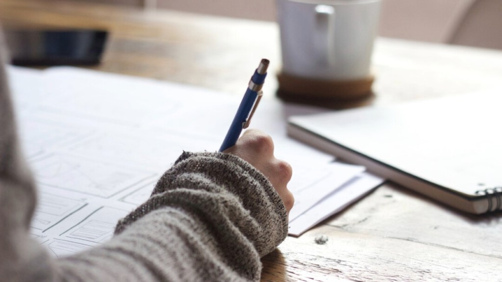 A person's hand holding a pen, writing on a stack of papers on a wooden table, with a coffee cup and a notebook in the background.