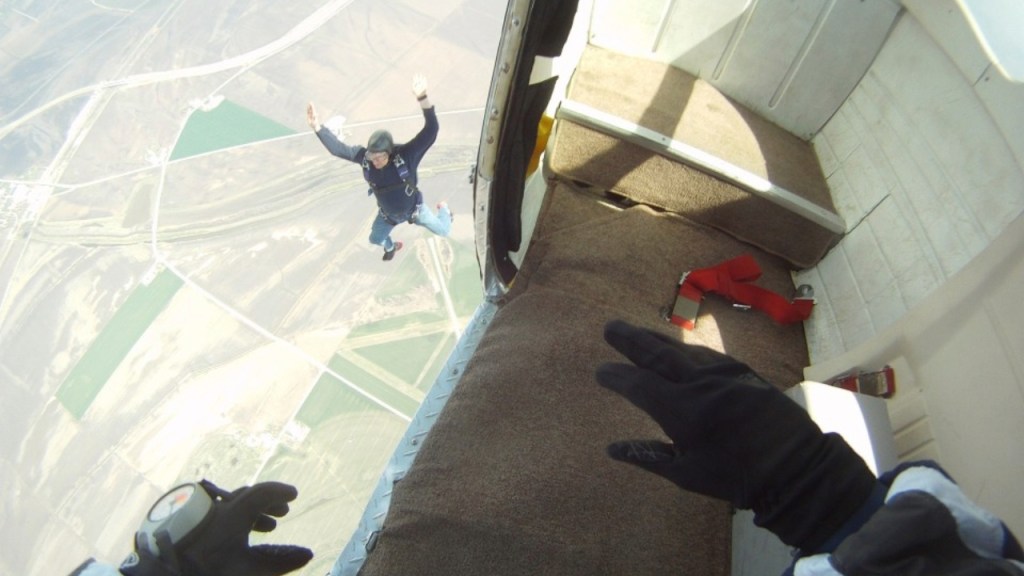 A skydiver jumping from an aircraft, arms raised, with the open side of the plane visible and a view of the landscape below.