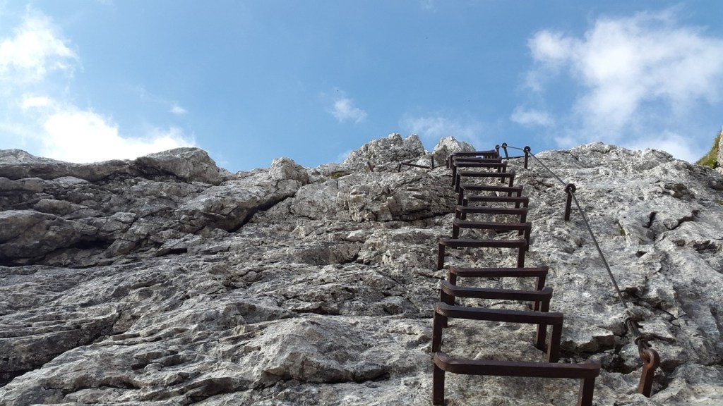 A metal ladder attached to a rocky cliff, leading up to a blue sky with scattered clouds.