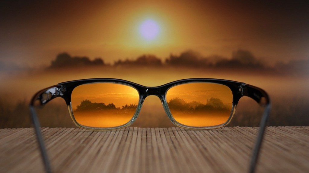 A pair of sunglasses with reflective orange lenses displaying a sunset landscape in the reflection, set against a soft-focus background of a hazy sunset.
