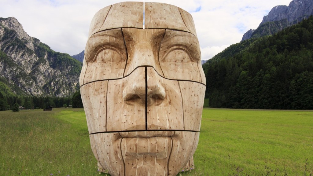 A large wooden sculpture resembling a human face, situated in a green field with mountains in the background. The sculpture is divided into sections, highlighting its intricate design against a cloudy sky.
