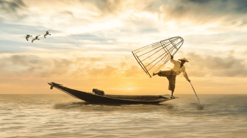 A fisherman in traditional attire using a net on a wooden boat during a sunset, with birds flying in the background.
