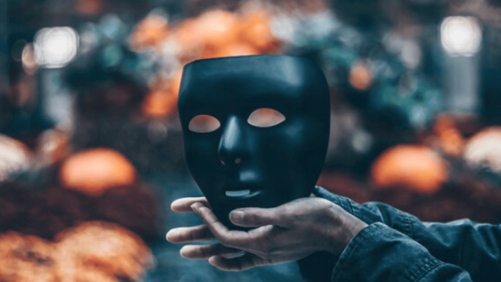 A hand holding a black mask against a blurred background with autumn decorations and pumpkins.