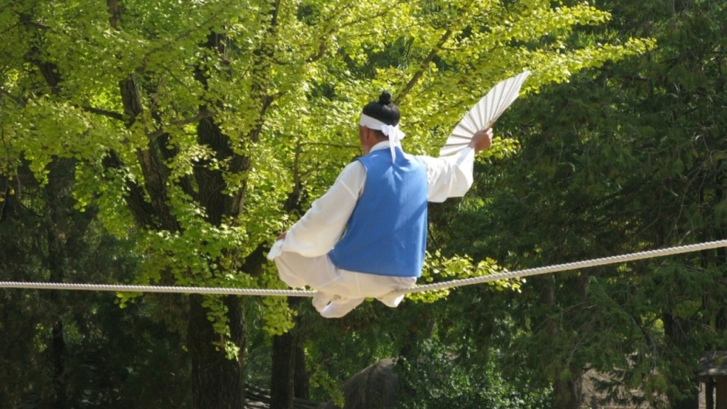 A performer dressed in traditional attire, balancing on a tightrope while holding a fan, with lush green trees in the background.